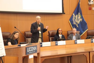 A speaker addresses an audience at a conference panel held at UConn Law. Five other panelists are seated at the long table, with name placards in front of them and the Connecticut state flag in the background.