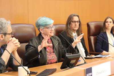 A panelist gestures while speaking during a conference discussion. Other panelists, including a man and a woman in a navy blazer, listen attentively.