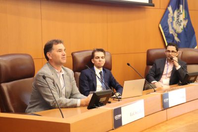 Three panelists, including a man in a gray blazer speaking into a microphone, participate in a discussion. A laptop and microphones are set up in front of them, and the Connecticut state flag is visible behind them.