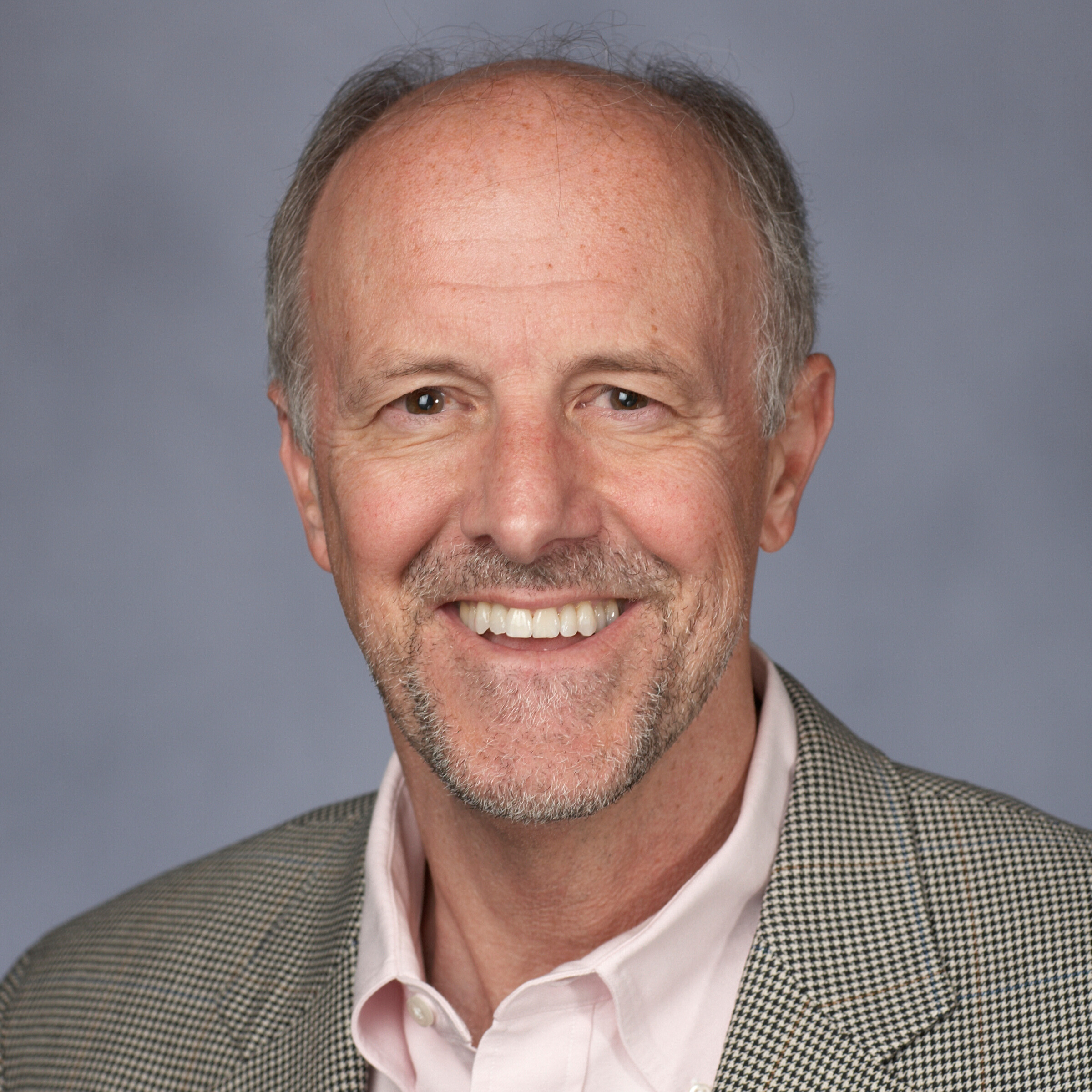Headshot of a man wearing a light pink collared shirt and patterned grey suit jacket