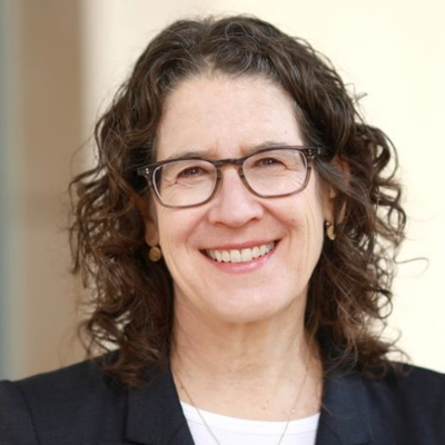 Professional headshot of a smiling person with shoulder-length curly dark hair, glasses, and a dark blazer against a softly blurred outdoor background.