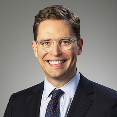 Professional headshot of a smiling person with glasses, wearing a dark suit, light blue shirt, and patterned tie against a gray background.