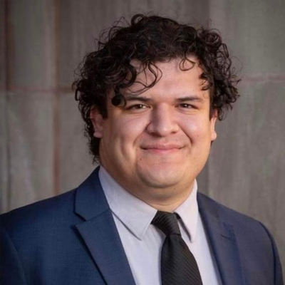 Professional headshot of a smiling person with curly dark hair, wearing a blue suit, gray shirt, and black tie against an outdoor background.
