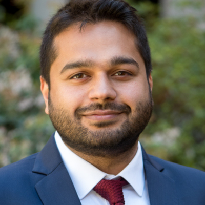 Professional outdoor headshot of a person with short dark hair and a beard, wearing a navy suit, white shirt, and burgundy tie in front of a blurred green background.
