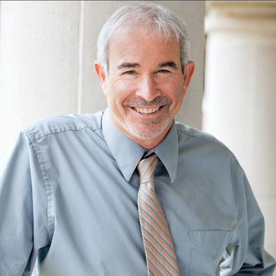 Professional headshot of a smiling person with short gray hair, wearing a light blue dress shirt and striped tie, standing outdoors against a column.