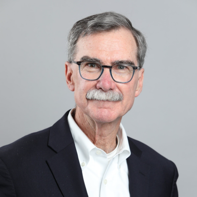 Professional headshot of a person with gray hair, glasses, and a mustache, wearing a dark blazer and white shirt against a gray background.