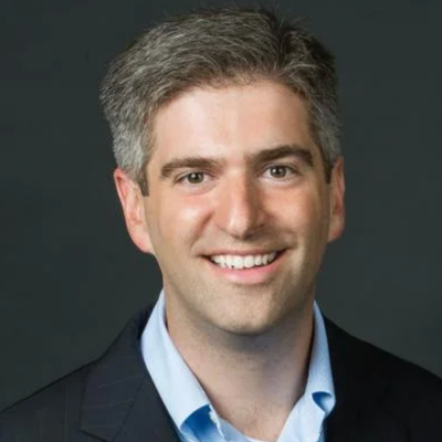 Professional headshot of a smiling person with short salt-and-pepper hair, wearing a dark blazer and light blue shirt against a dark background.