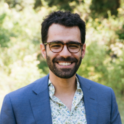 Professional outdoor headshot of a smiling person with dark hair, glasses, a beard, and a blue blazer over a patterned shirt.