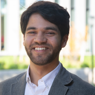 Professional outdoor headshot of a smiling person with dark hair and a short beard, wearing a gray blazer and white shirt.