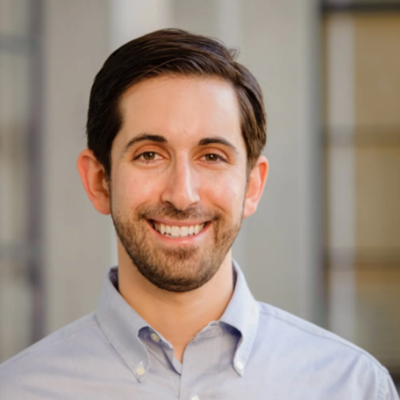 Professional headshot of a smiling person with short dark hair, wearing a light blue button-down shirt in front of a softly blurred building.