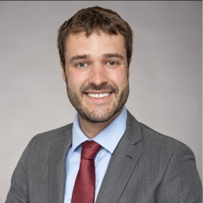 Professional headshot of a smiling person with short brown hair and light facial hair, wearing a gray suit, light blue shirt, and burgundy tie against a gray background.